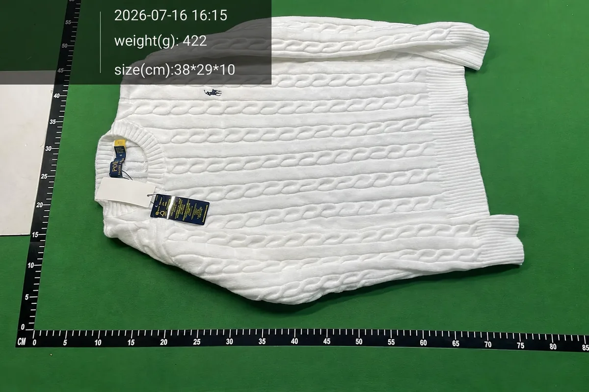 White cable-knit jumper laid flat on a green warehouse inspection surface with a measurement scale